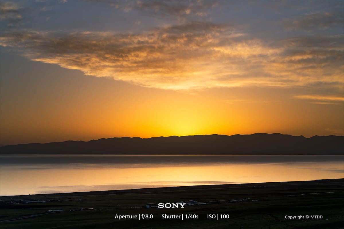 Golden sunset over mountain silhouettes reflects on calm lake waters, creating a serene landscape with dramatic sky