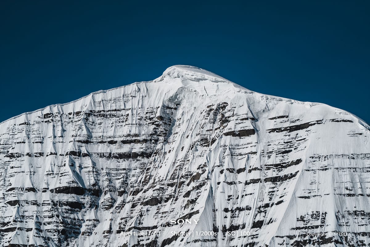 Snow-covered mountain peak with steep rocky cliffs and glacial formations against deep blue sky