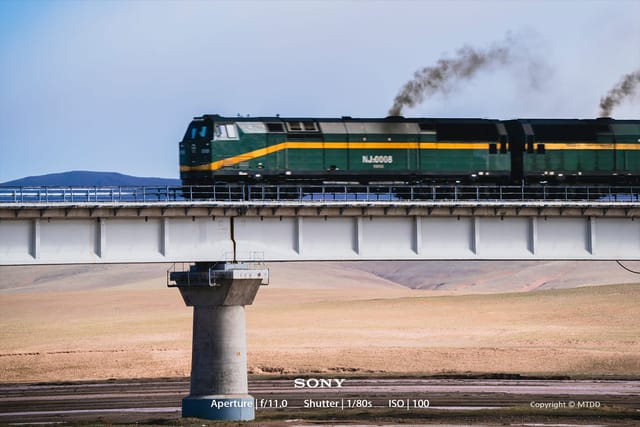 Green and yellow locomotive crossing elevated railway bridge over salt flats with mountains in background under blue sky