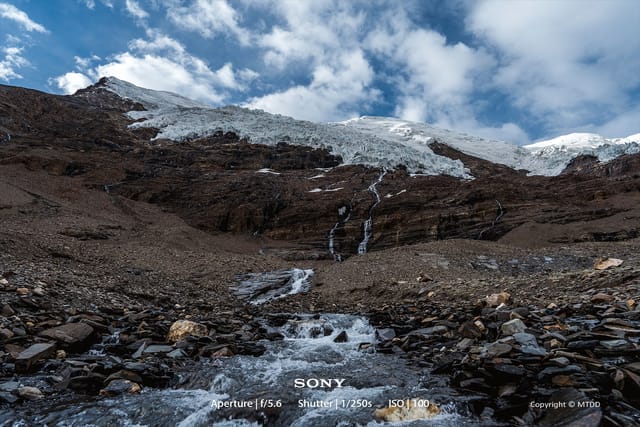 Snow-capped mountain peak with cascading glacial waterfalls flowing over rocky terrain under dramatic cloudy sky