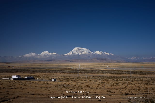 Snow-capped Himalayan mountain peak rises above vast Tibetan plateau with scattered buildings and power lines across arid landscape